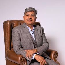 Narsi Kularia in a grey three-piece suit, seated in a tan leather armchair against a pale wall, smiling towards the camera