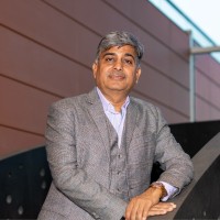 Narsi Kularia in a grey checked blazer leaning on a dark railing before a terracotta-panelled building