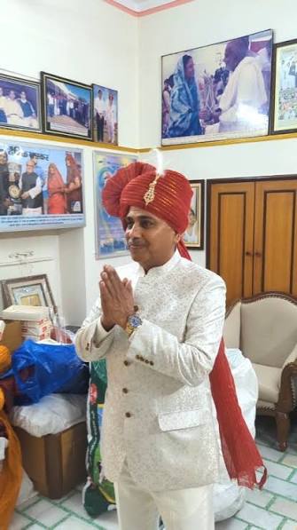 Narsi Kularia in a red Rajasthani safa and ivory sherwani, hands folded in greeting, in a room lined with framed photographs